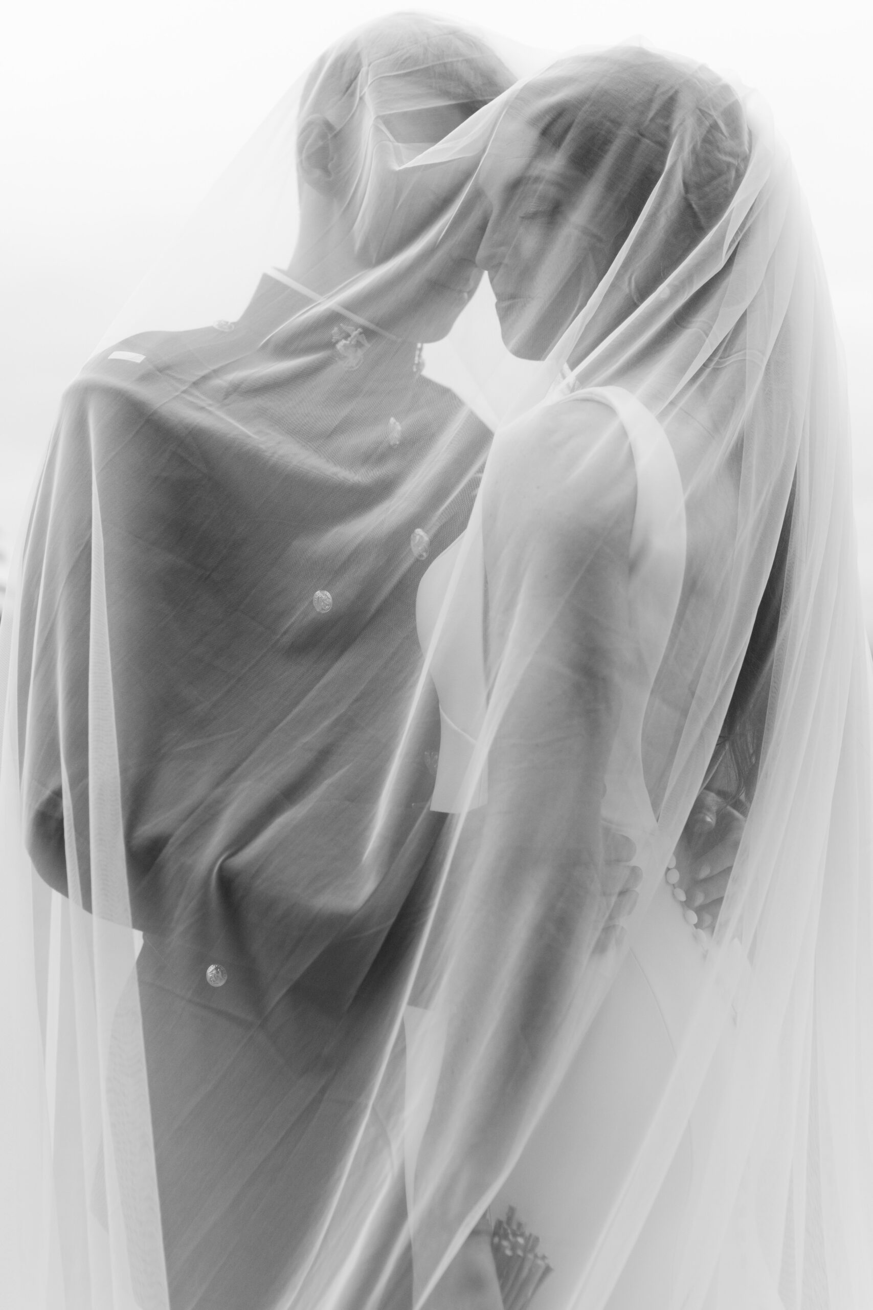 Black and white photo of bride and groom embracing covered in her veil.